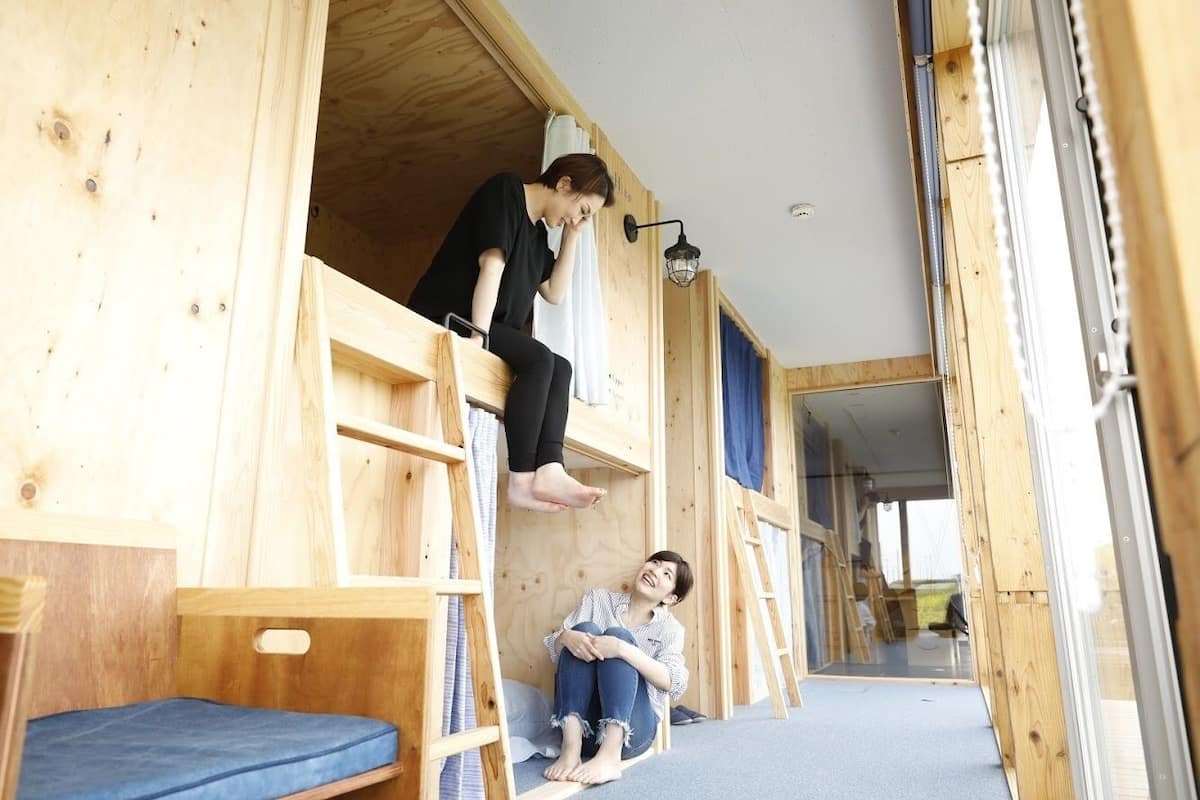 A wooden interior with a loft bed and a lower bunk bed, a ladder, and a small blue mattress on the floor.