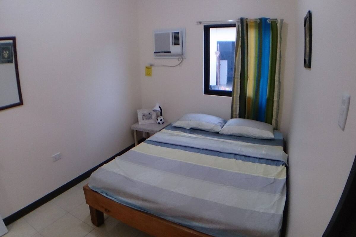 A small, single-bed room with a striped bedspread, a bedside table with a lamp, and a window with curtains.