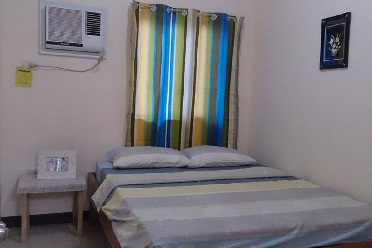 A single bed with a striped bedspread, a bedside table with a lamp, an air conditioning unit, and a curtain with vertical stripes.