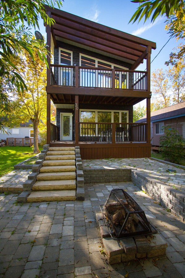 A two-story wooden house with a balcony, a fire pit, and a paved driveway.