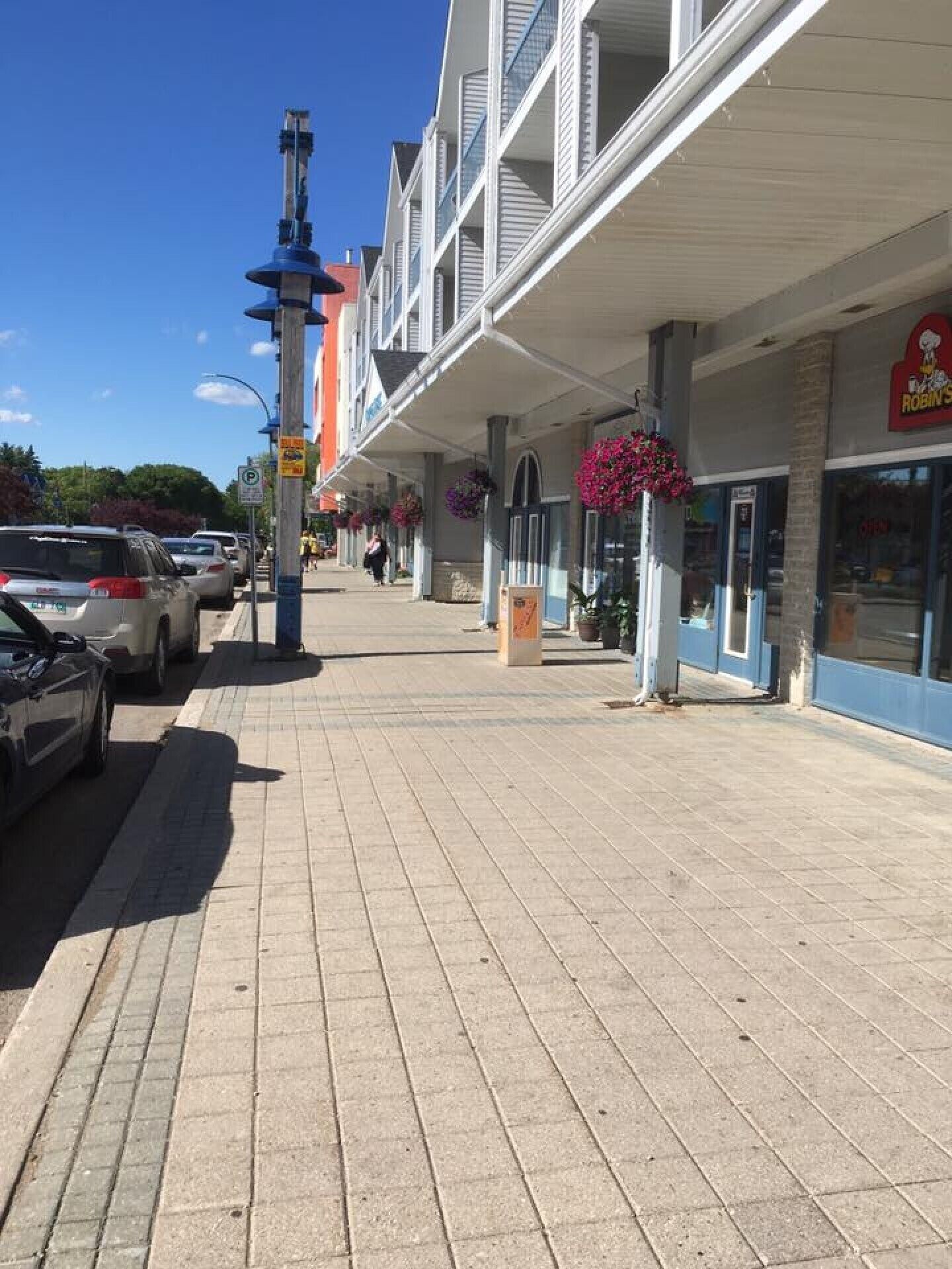 A sidewalk with parked cars, a pedestrian walkway, and storefronts with hanging flower baskets.