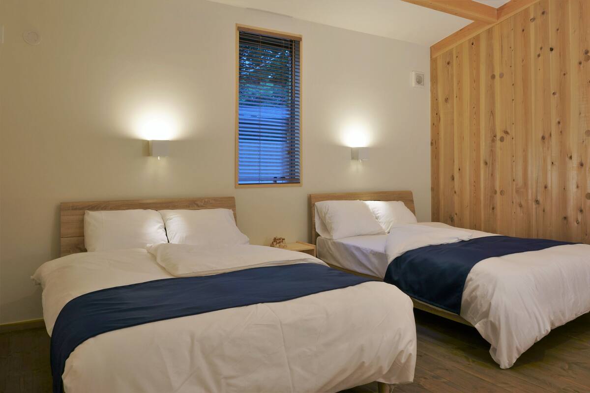 A room with two beds, wooden walls, a window with blinds, and wall-mounted lights.