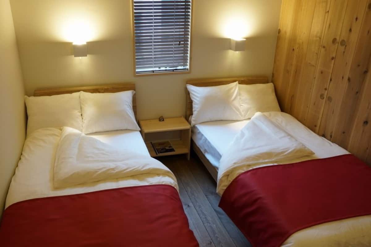A small, cozy room with two beds, each with white bedding and red accents, wooden headboards, and a small bedside table between them.