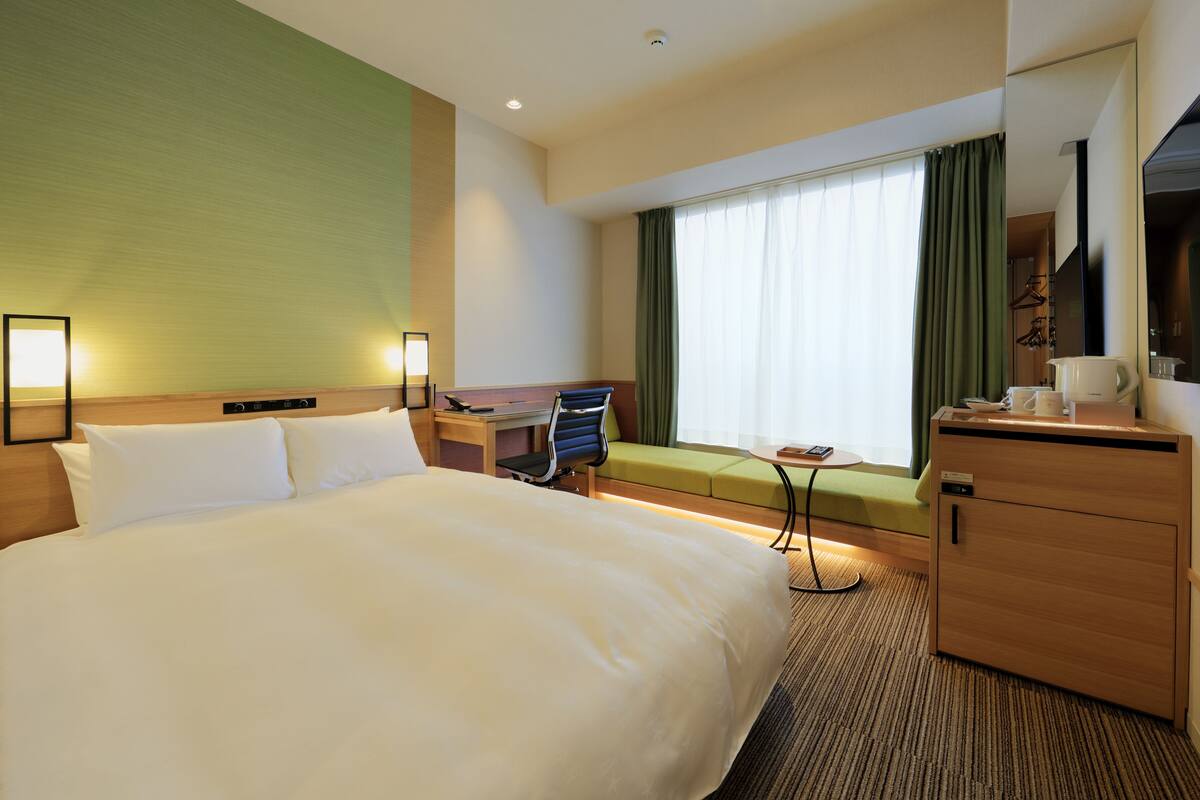 A hotel room with a large bed, a desk, a chair, a small table, a cabinet, and a window with curtains.