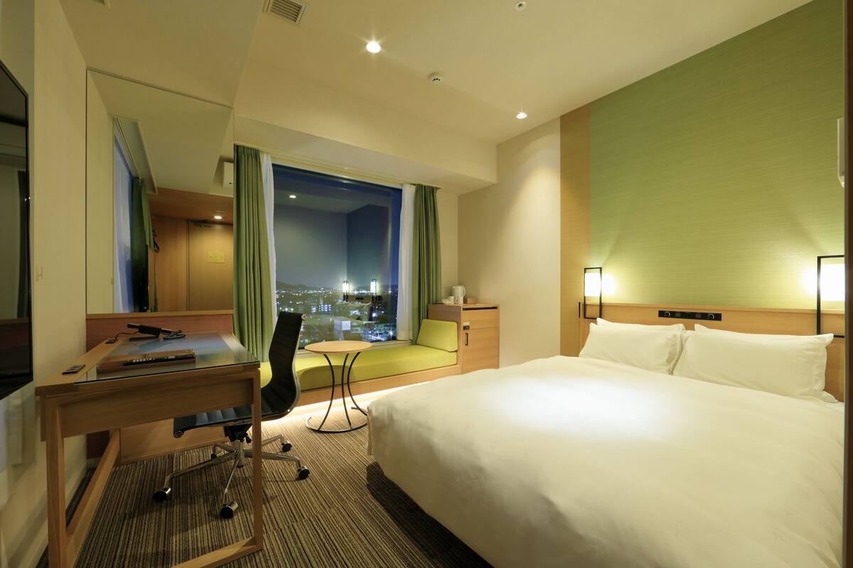 A hotel room with a bed, desk, chair, and a view of the city at night.