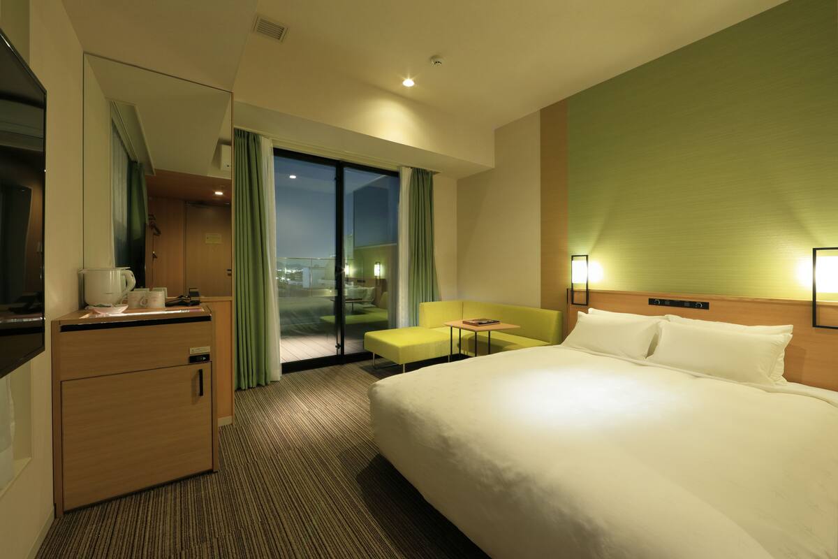A hotel room with a bed, a desk, a chair, a small table, and a view of the city at night.