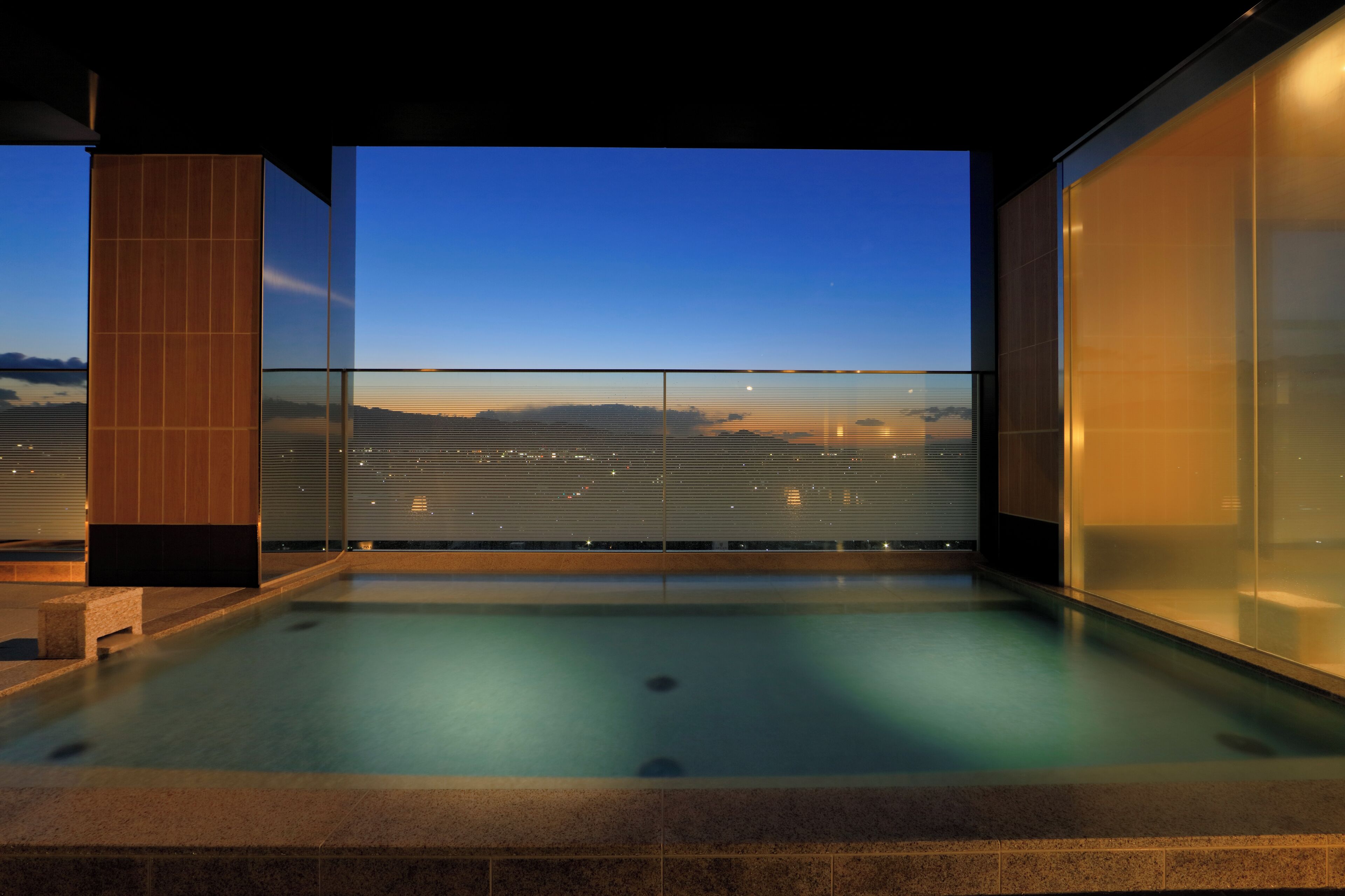 A modern indoor pool with a glass barrier and a view of the cityscape at dusk.