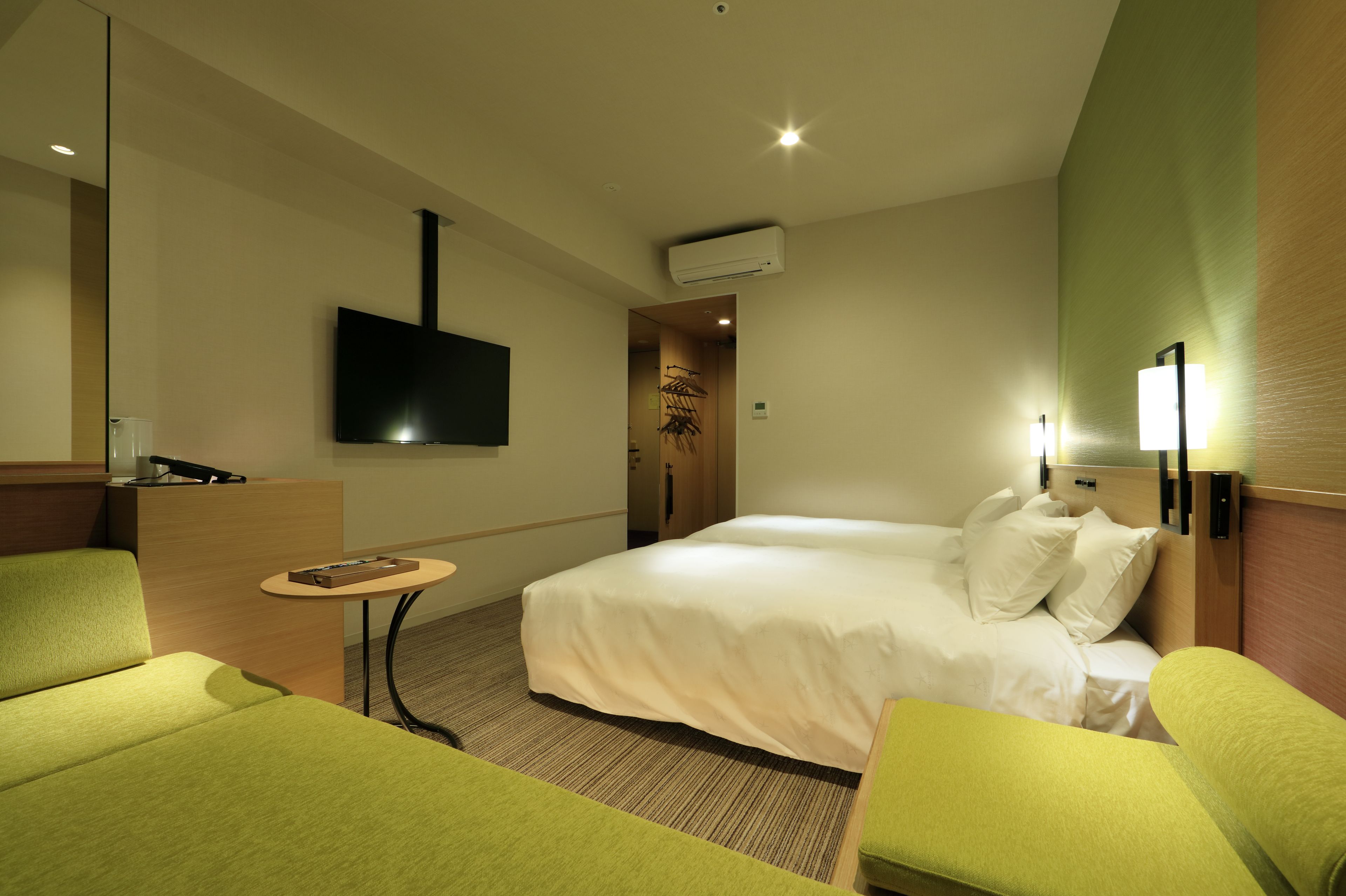 Une chambre d’hôtel avec un lit, deux fauteuils verts, une petite table, un téléviseur fixé au mur et un luminaire fixé au mur.