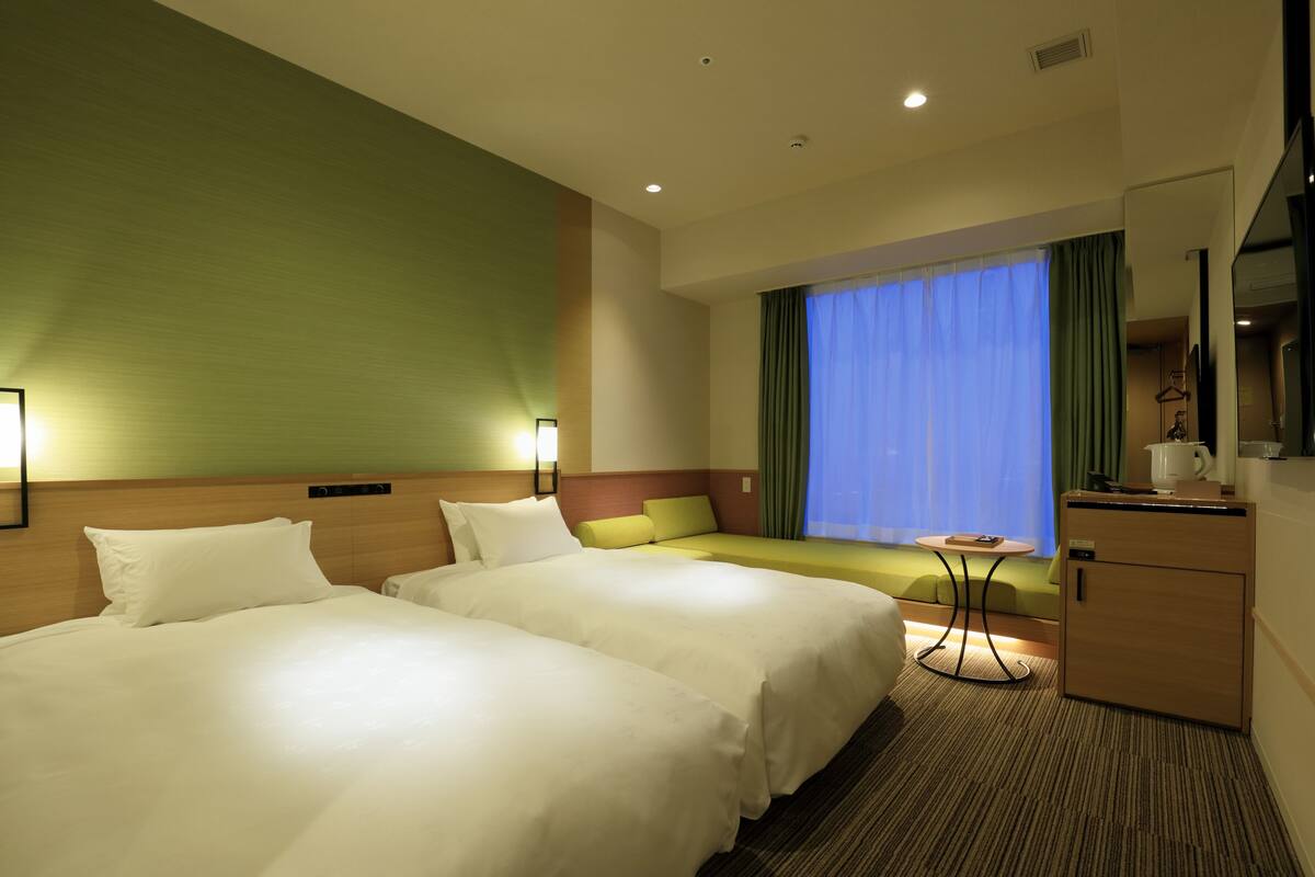 A hotel room with two beds, a small table, a sofa, a desk, and a window with curtains.
