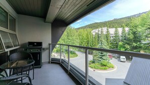Ein Balkon mit Grill, Korbstühlen und Blick auf einen Parkplatz sowie bewaldete Berge.