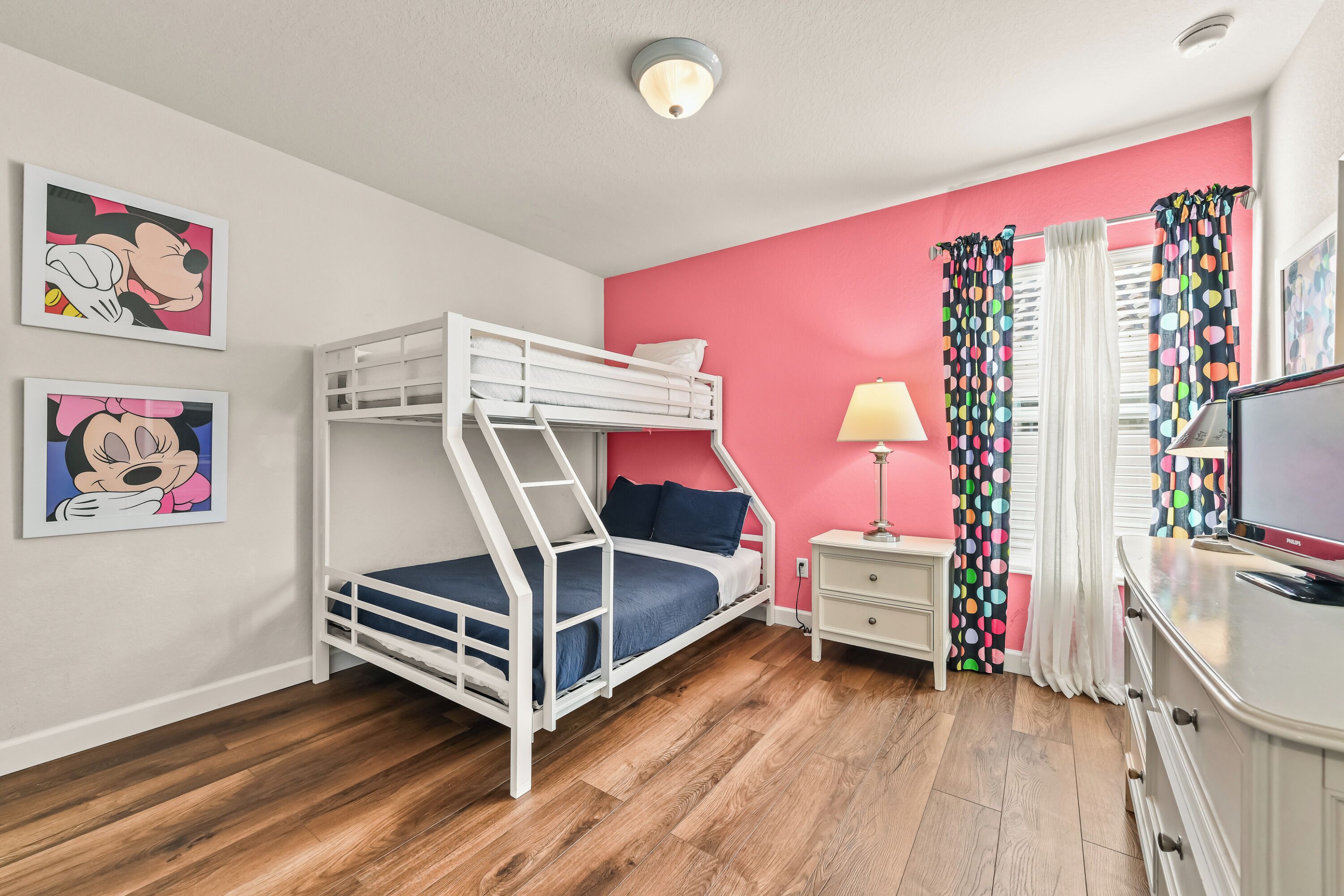Ein Schlafraum mit Etagenbetten, einer rosafarbenen Wand, Holzboden, einem Fernseher, einer Lampe und Mickey Mouse Postern.