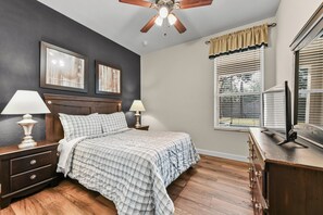 A bedroom with a bed, two bedside tables with lamps, a ceiling fan, a window with blinds, and a dresser.