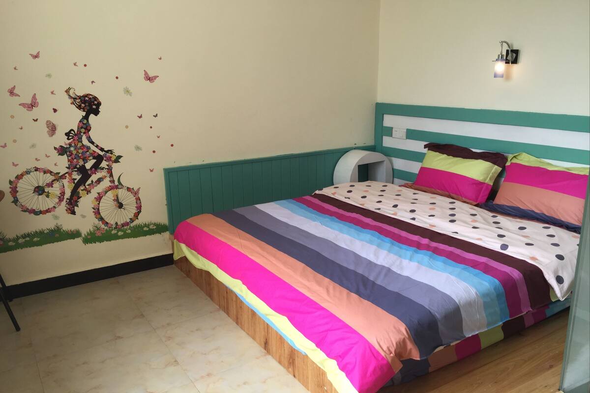 A bedroom with a bed, colorful bedding, a wall mural, and a nightstand.