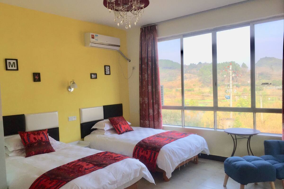 A hotel room with two beds, a small table, and a view of a landscape through large windows.