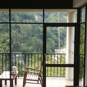 A balcony with a wooden table, a chair, and a view of a lush green landscape with a waterfall and buildings in the distance. - Viewfront Ella (Ella)