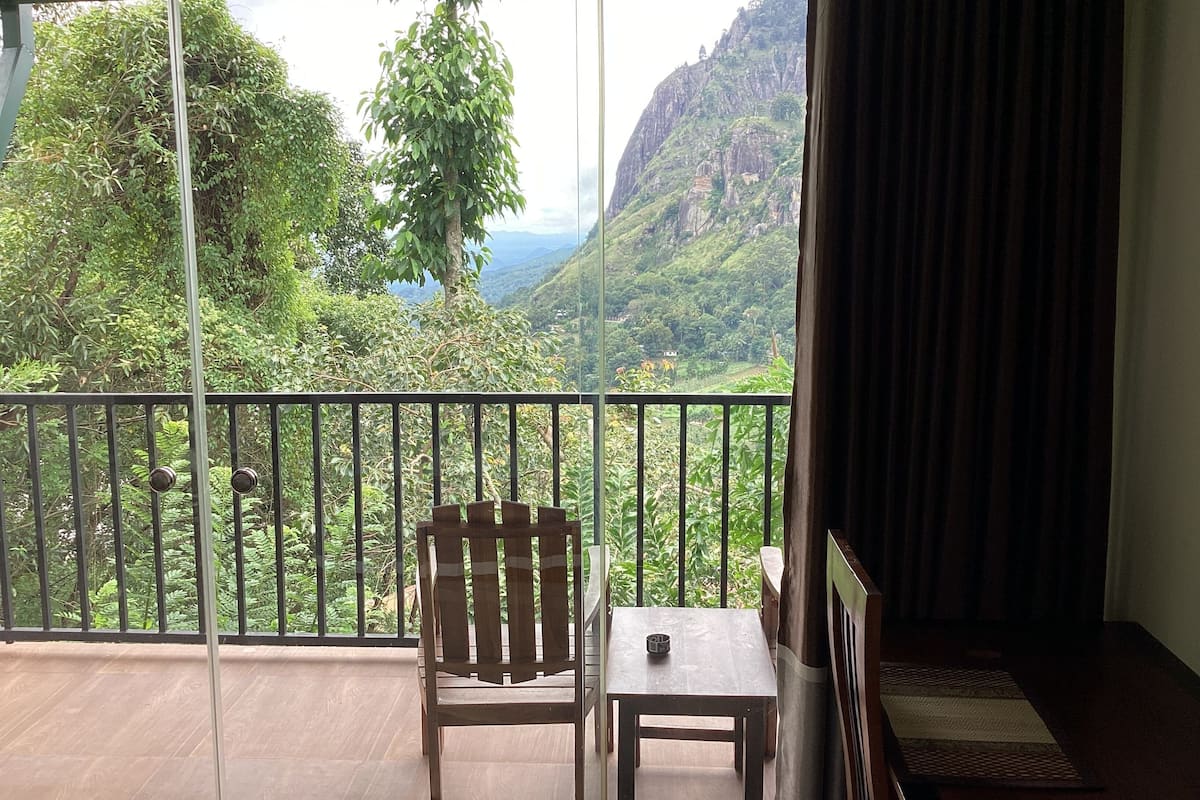 Uma varanda com cadeira e mesa de madeira, com vista para um vale exuberante com montanhas e vegetação.