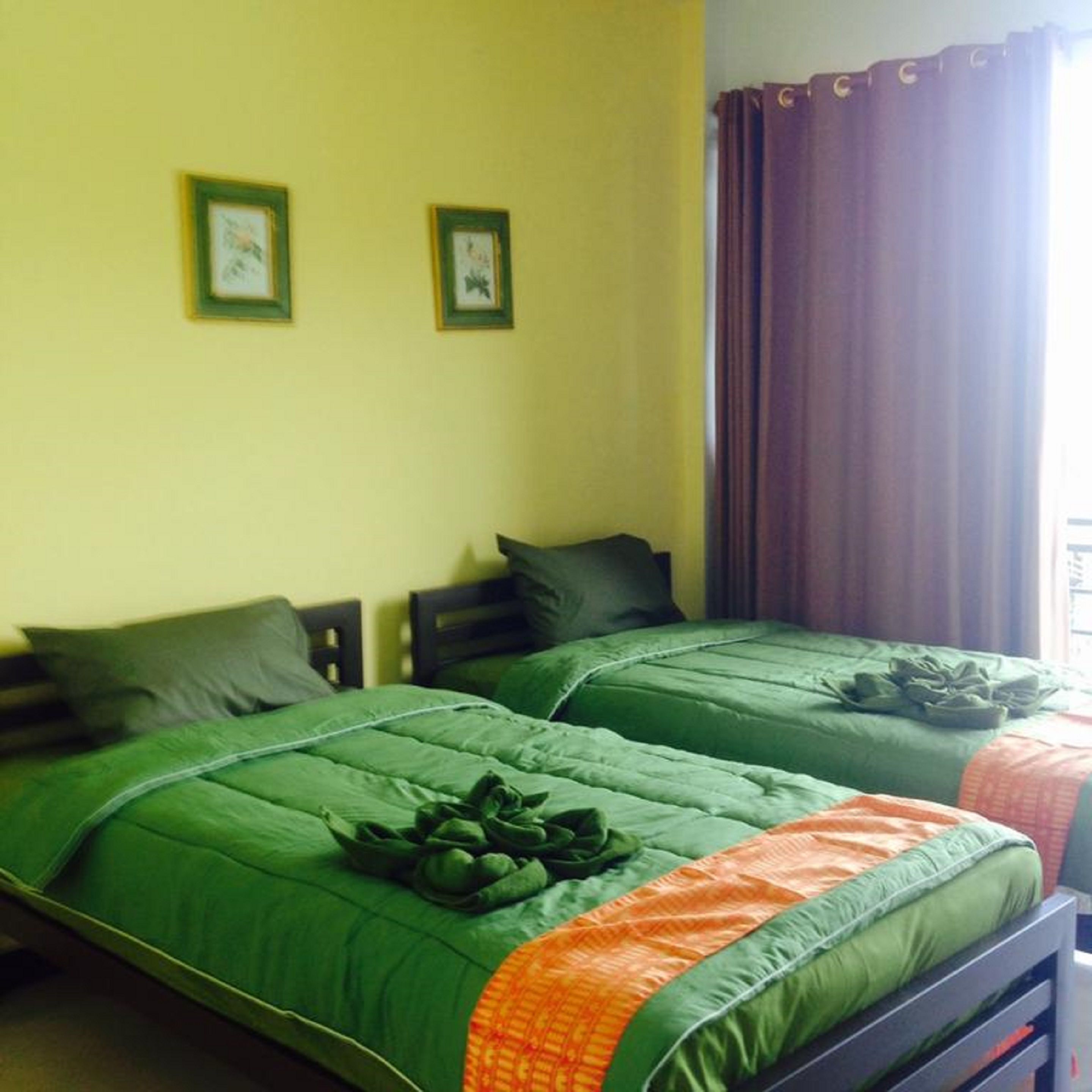 Quarto com duas camas de solteiro, cada uma com roupa de cama verde e um travesseiro decorativo. Há duas imagens emolduradas na parede.