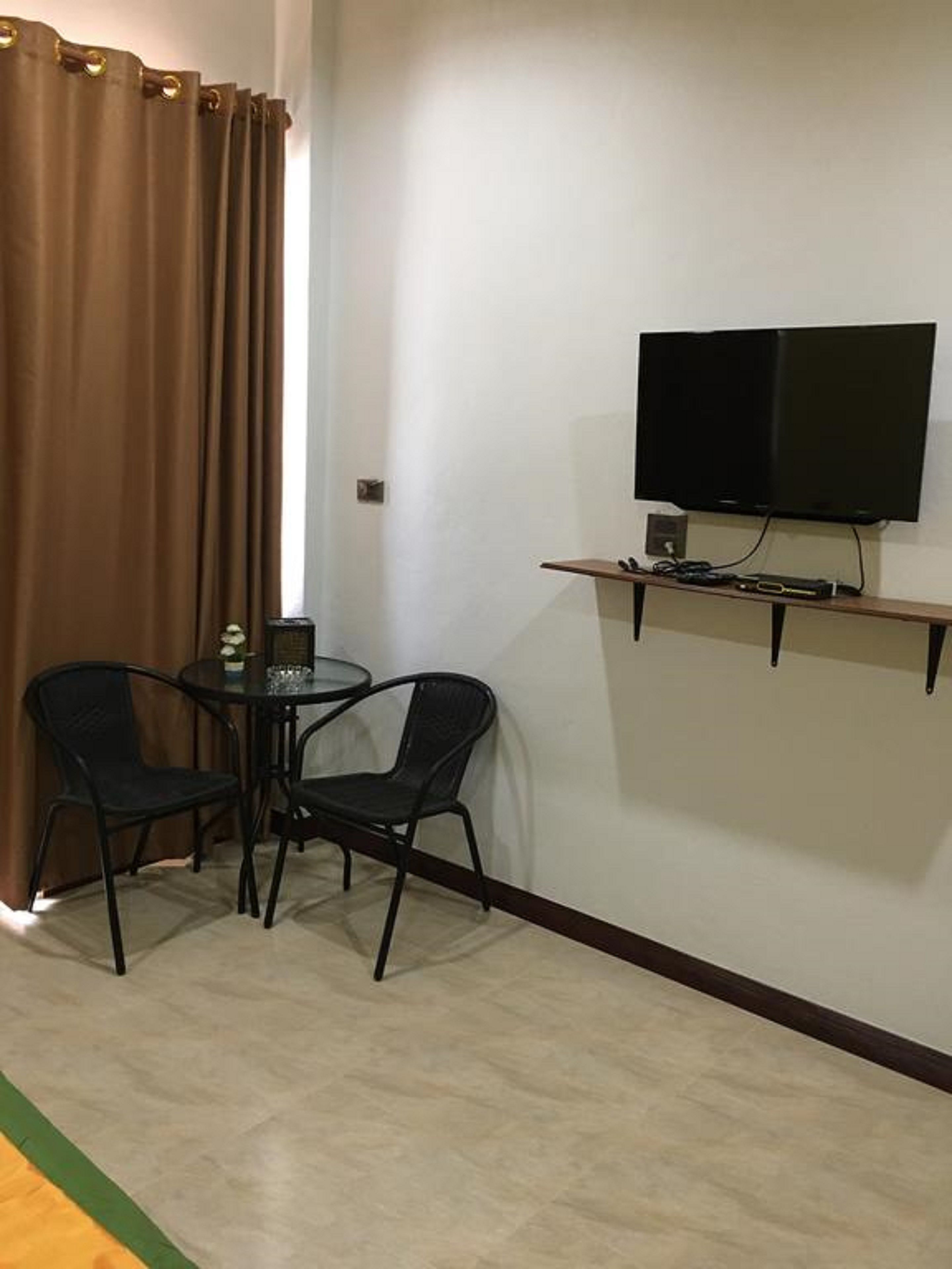 Een kleine kamer met een aan de muur bevestigde televisie, twee stoelen, een klein tafeltje en een gordijn.