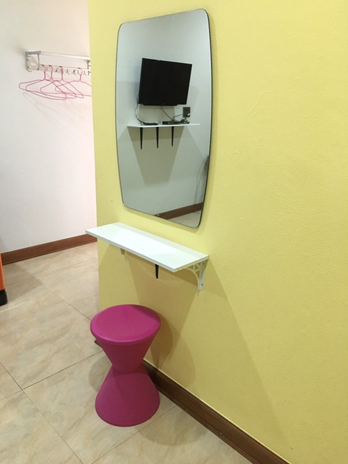 A bathroom with a yellow wall, a white shelf, a pink toilet, and a mirror reflecting a TV on a stand.