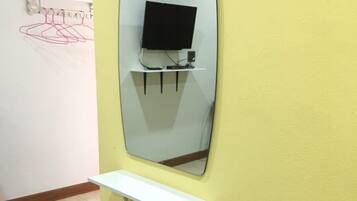 A bathroom with a yellow wall, a white shelf, a pink toilet, and a mirror reflecting a TV on a stand.