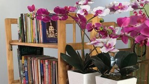 A bookshelf filled with books and two white planters with orchids. - Baan Suan Punpaka Resort (La-ngu)