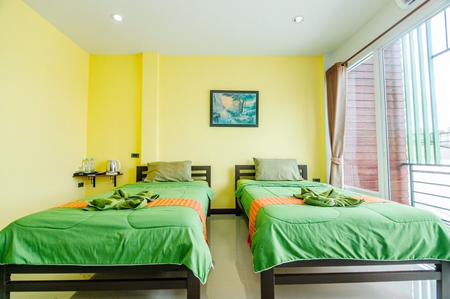 A room with two single beds, each with green bedding and a pillow. There is a painting on the wall, a window with curtains, and a small table with a water bottle and a kettle.