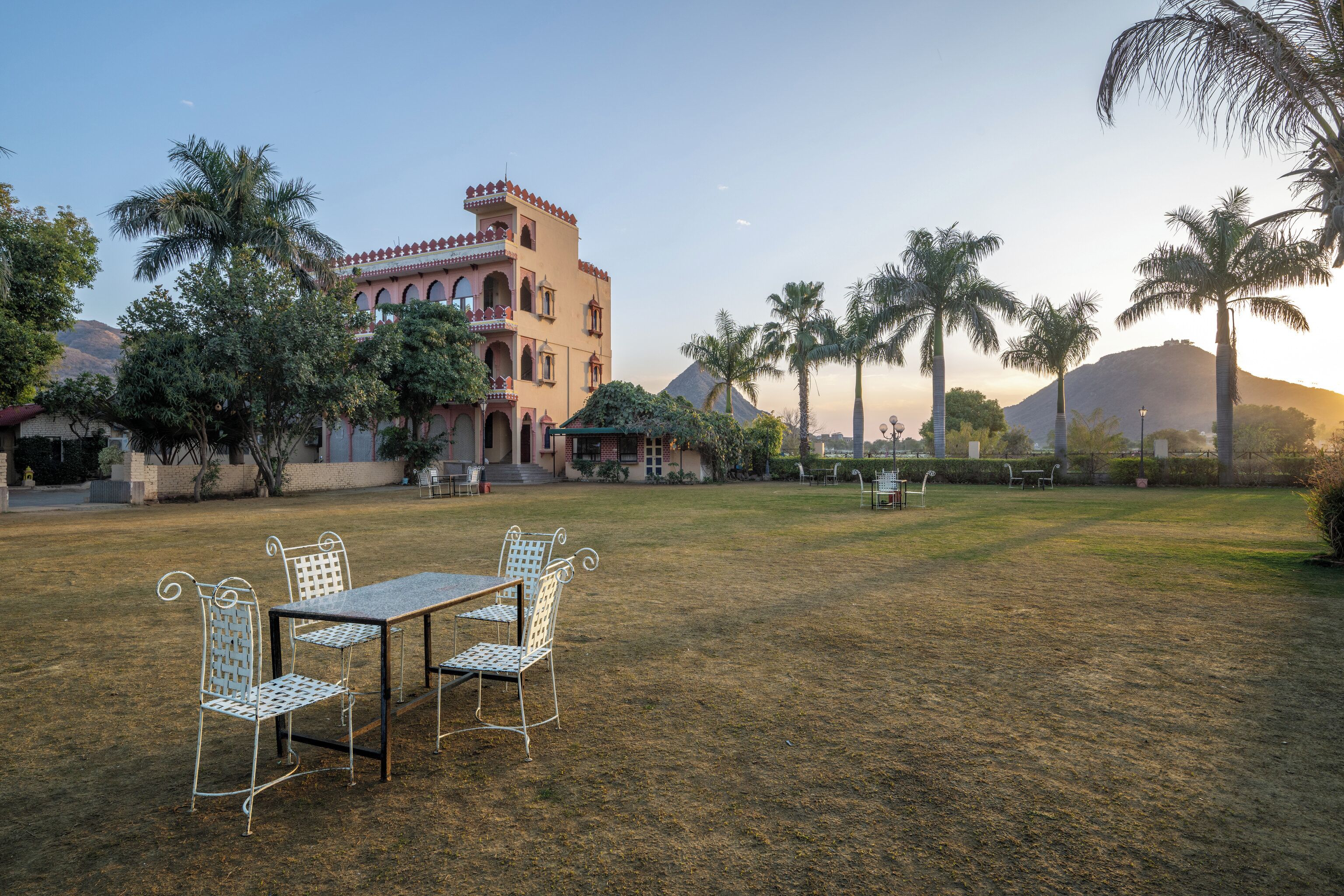 A courtyard with a table and chairs, palm trees, and a building with arched windows and a tower.