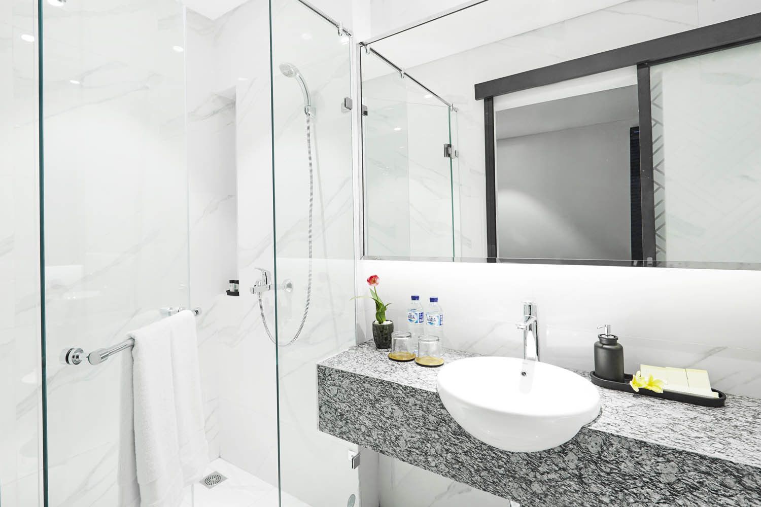 Un baño moderno con ducha envuelta en vidrio, encimera de granito, lavamanos blanco y espejo sobre el lavamanos.