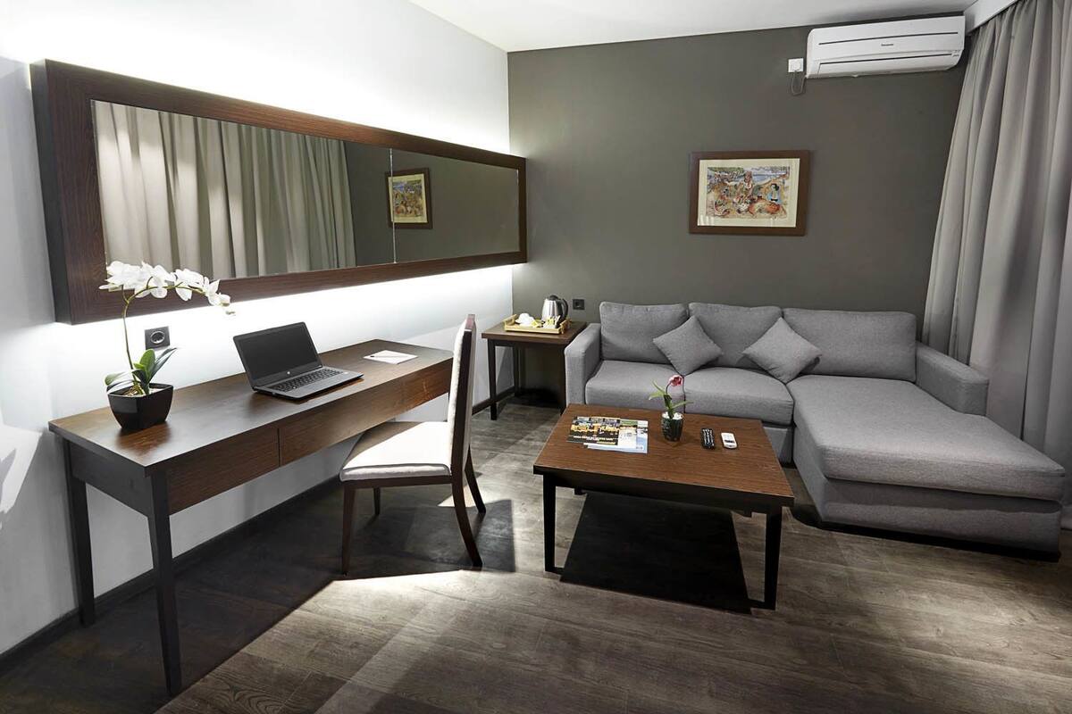 A modern living room with a grey sofa, a wooden desk with a laptop, a coffee table with magazines, and a potted orchid.