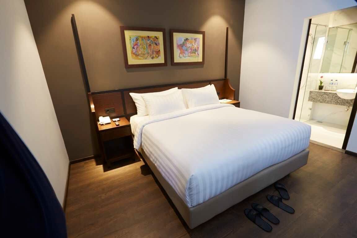 A modern hotel room with a large bed, wooden flooring, a nightstand, and a bathroom visible through a glass door.