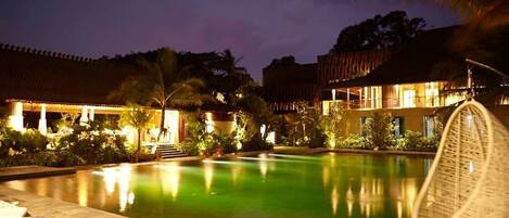 A nighttime view of a hotel pool area with a clear pool, lounge chairs, and a building with a tiled roof.