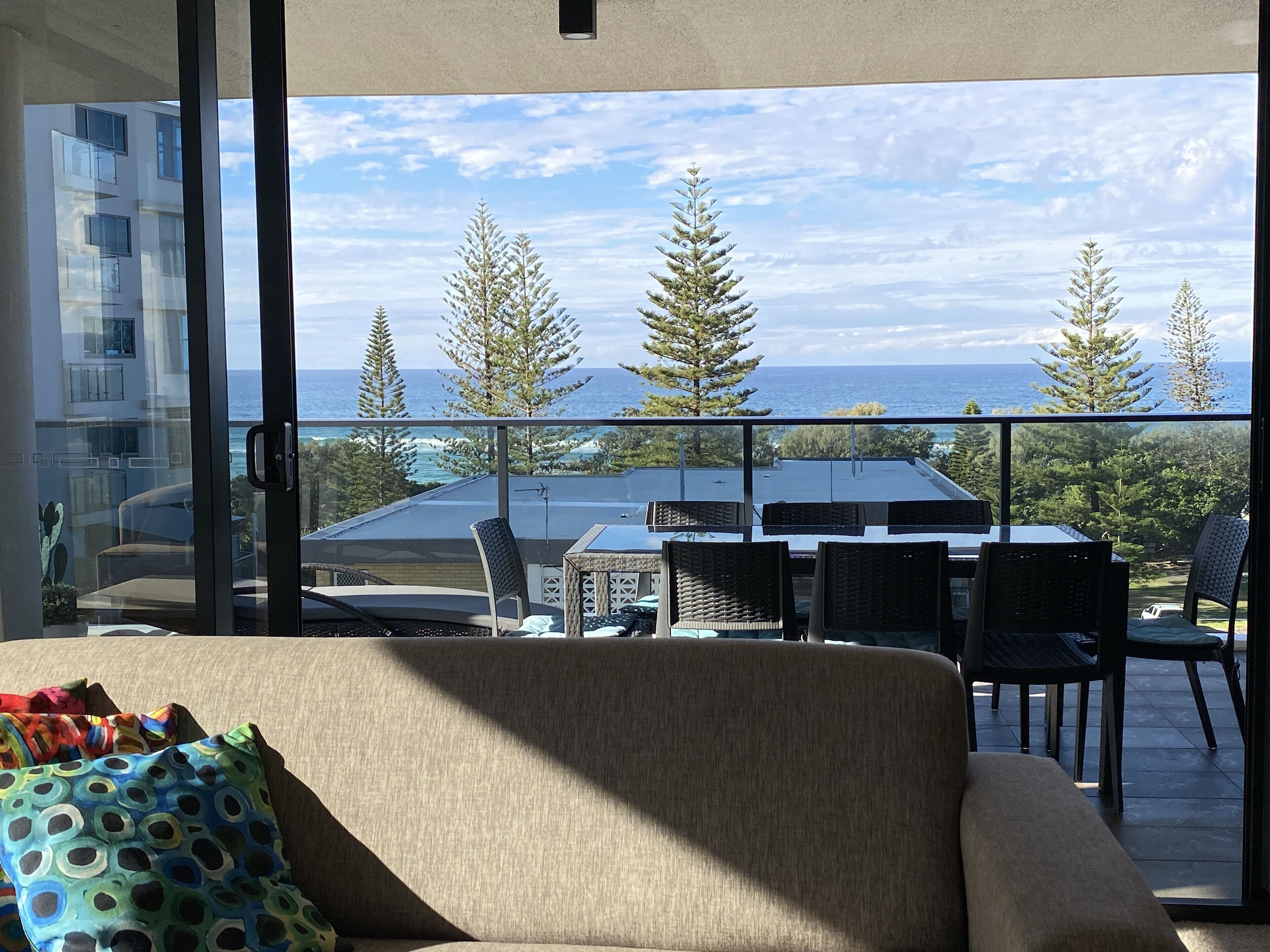 A balcony with a sofa and chairs, overlooking a coastal landscape with trees and a view of the sea.