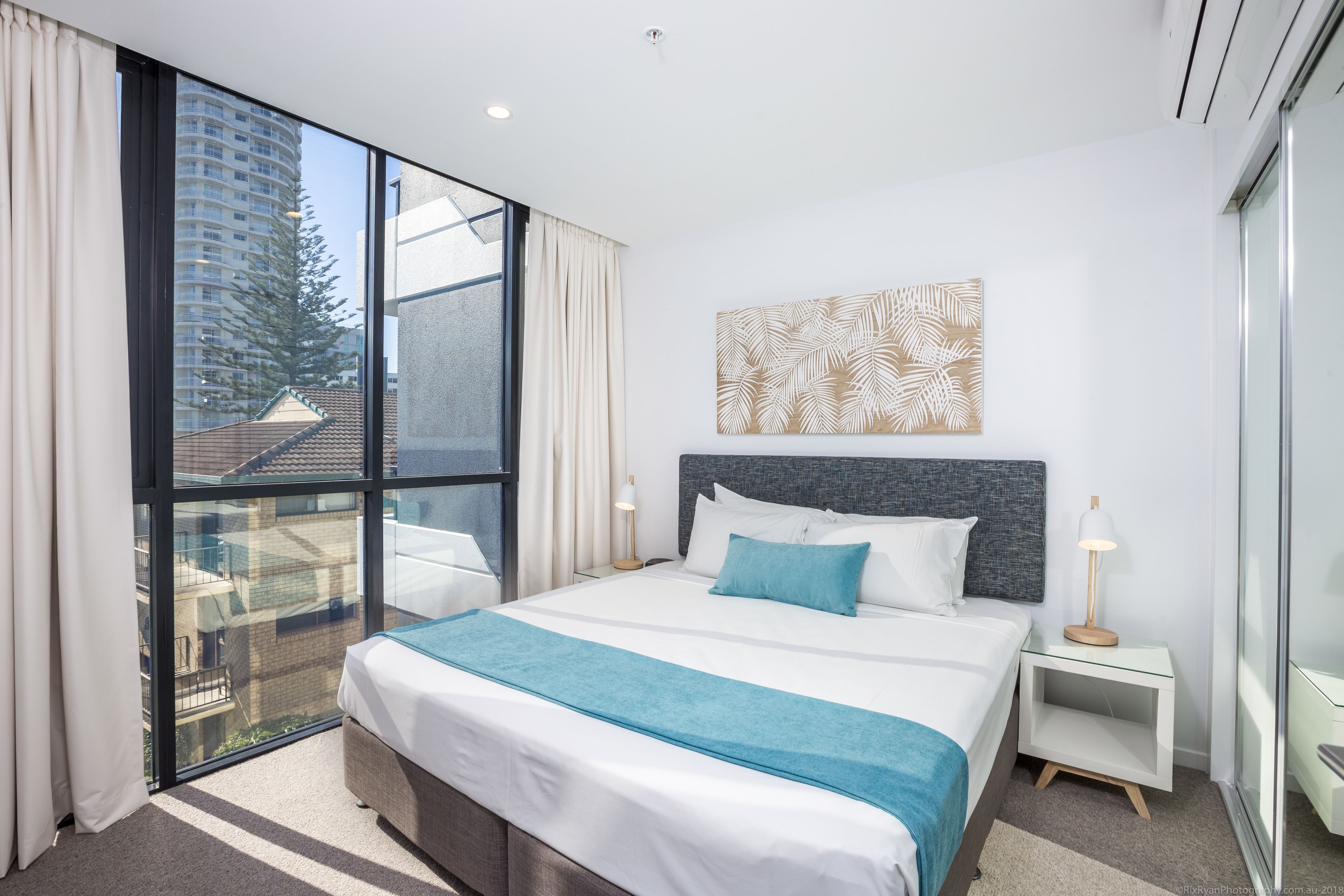 Habitación de hotel moderna con una cama grande, una mesita de noche, una lámpara de pared y un amplio ventanal con vistas a un edificio y un árbol.