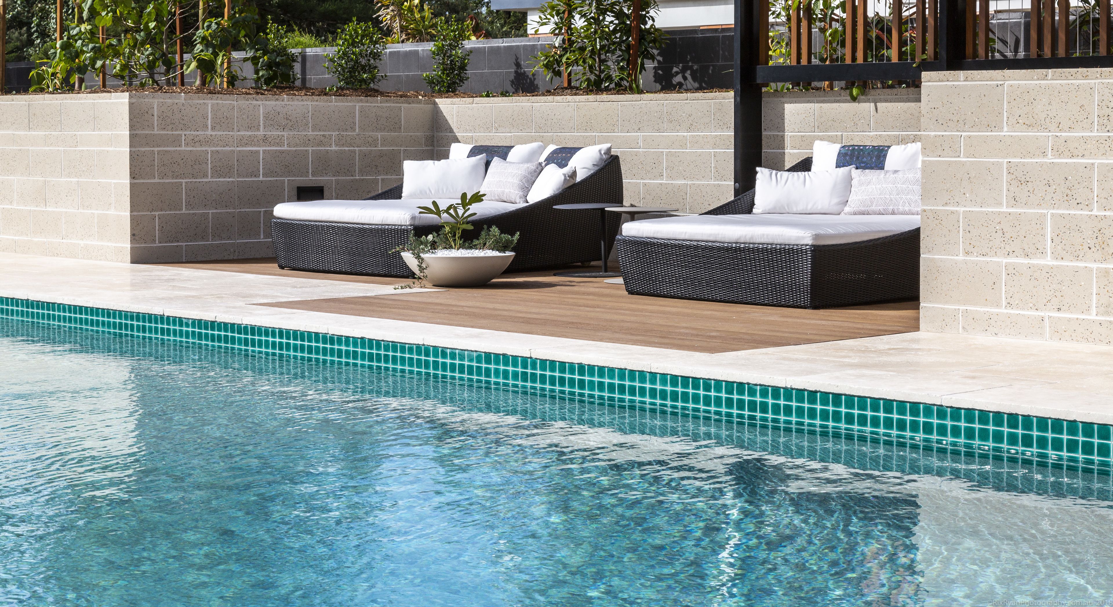A poolside area with two wicker lounge chairs, a potted plant, and a tiled pool edge.