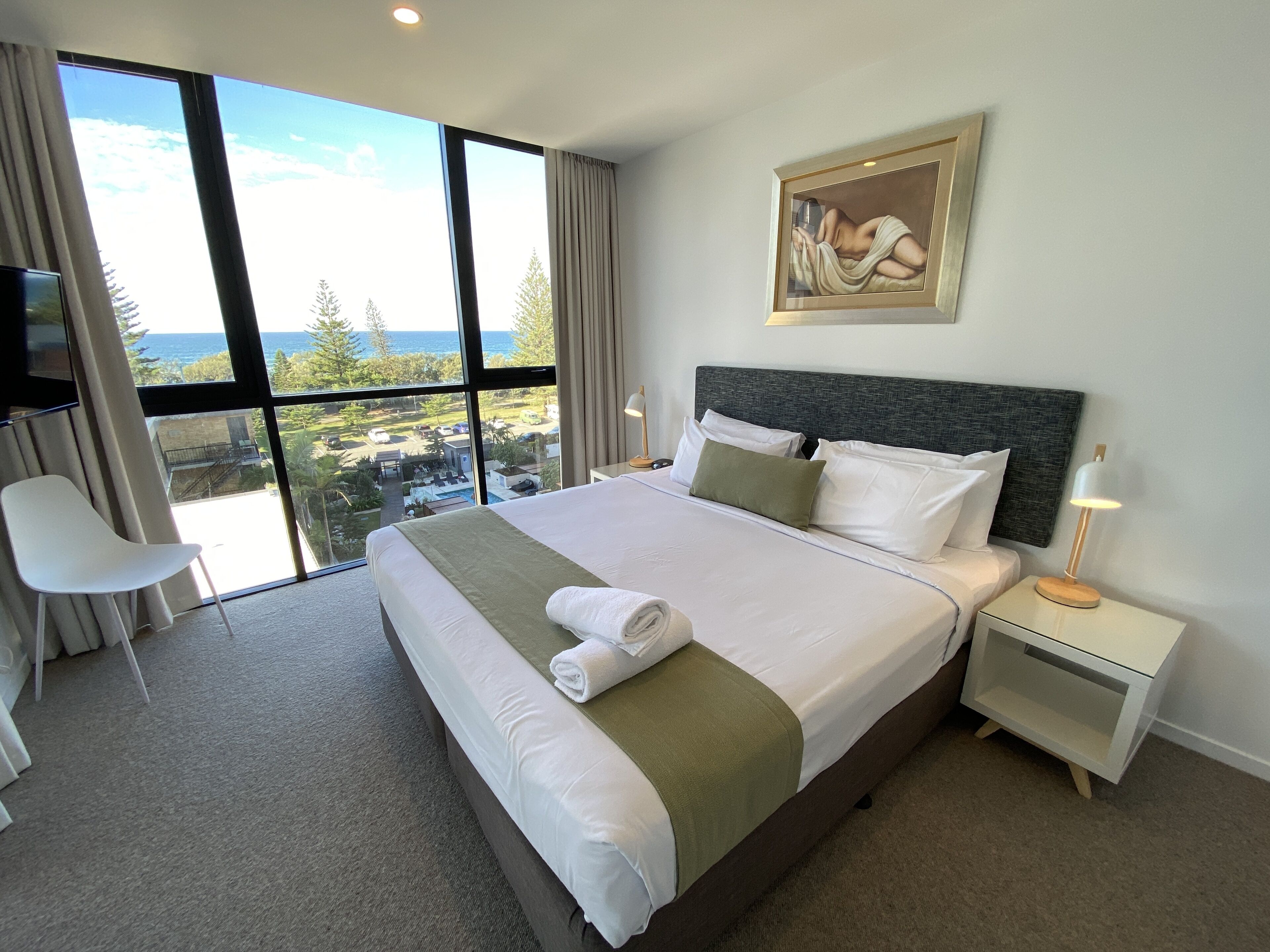 A hotel room with a large bed, a painting on the wall, a desk with a lamp, and a view of the sea through large windows.
