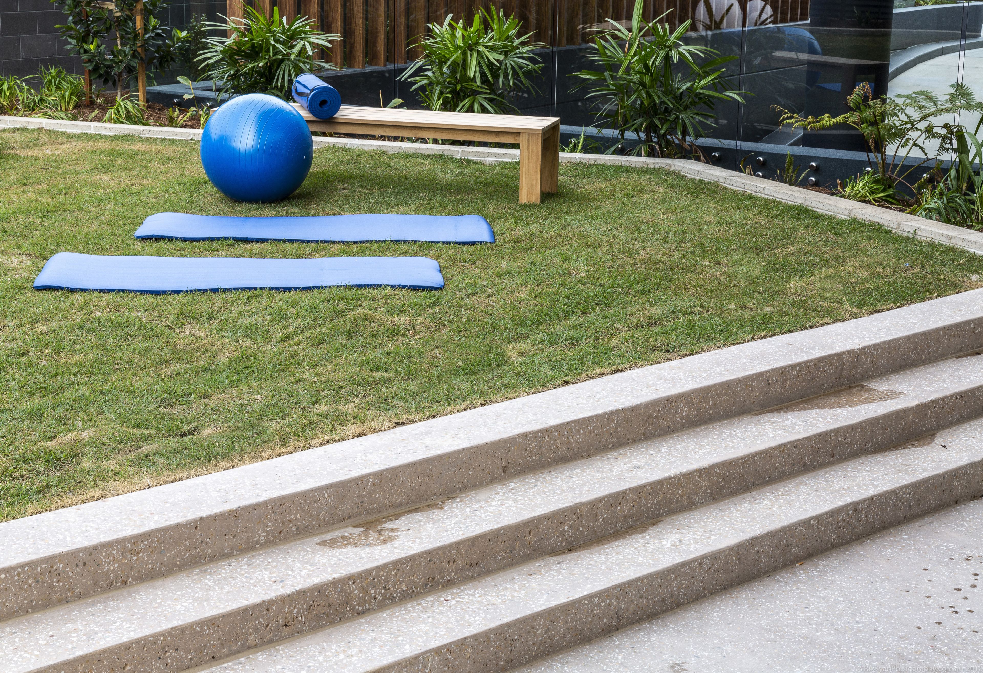 A grassy outdoor area with exercise equipment, including a large blue ball and yoga mats, and a wooden bench.
