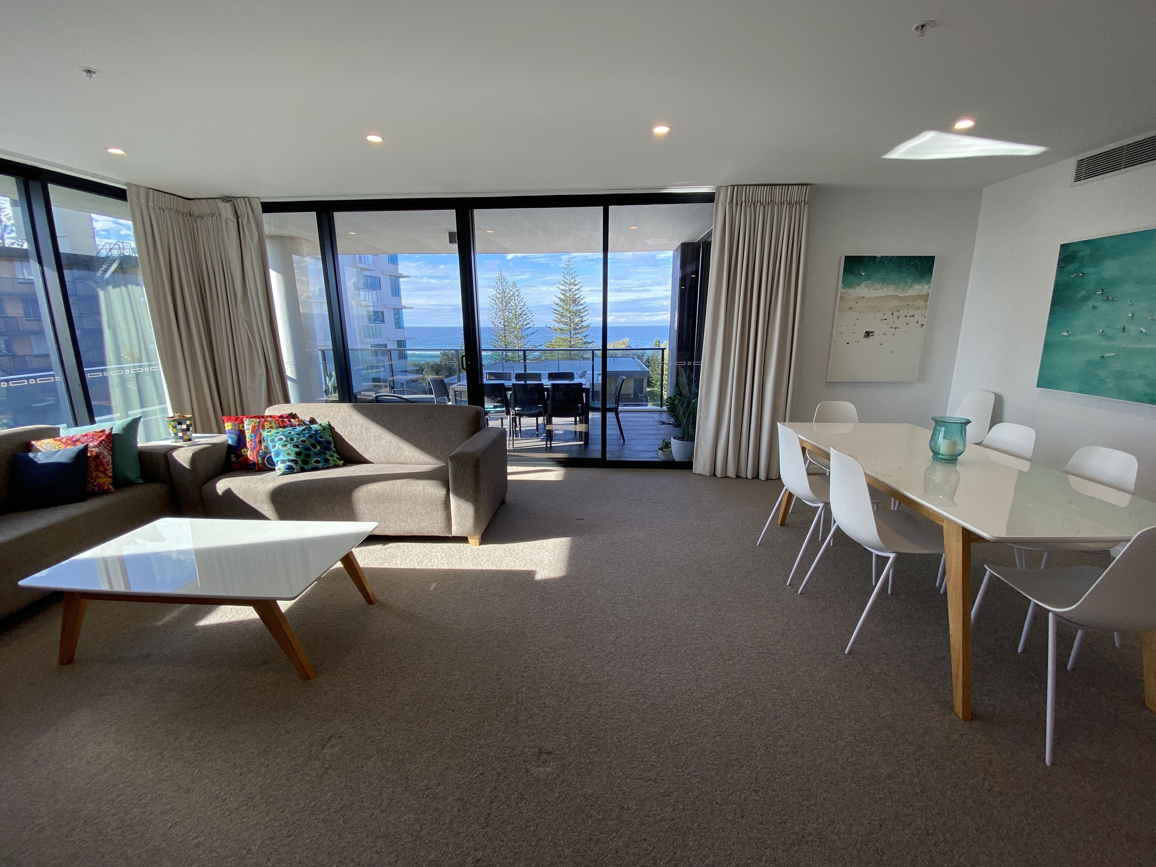 A modern living room with a sofa, coffee table, and dining table. Large windows offer a view of the city and a balcony with outdoor seating.