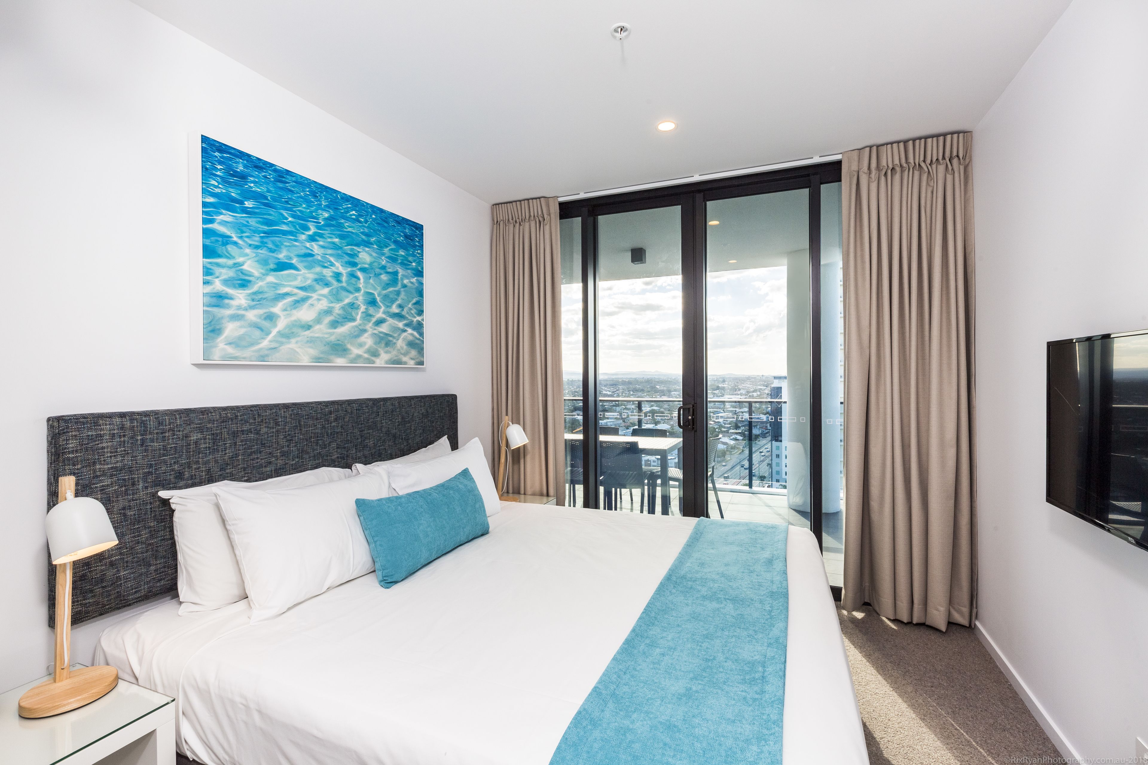 A modern hotel room with a large bed, a city view balcony, and a painting of water.