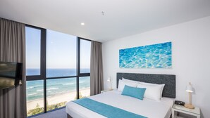 Habitación de hotel con un ventanal grande que da a la playa, una cama con ropa de cama blanca y azul, y un cuadro de agua en la pared.