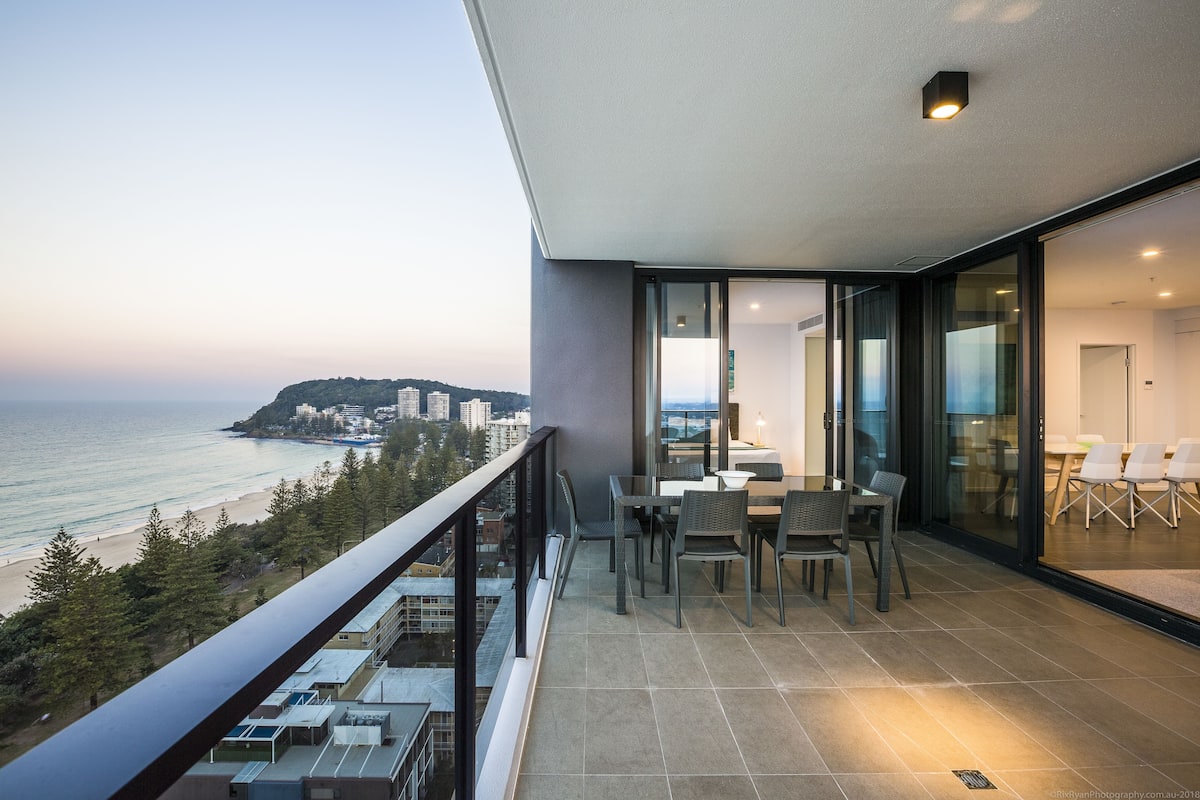 A balcony with a view of the sea and buildings, a dining area with chairs and a table, and a glass door leading to an interior space.