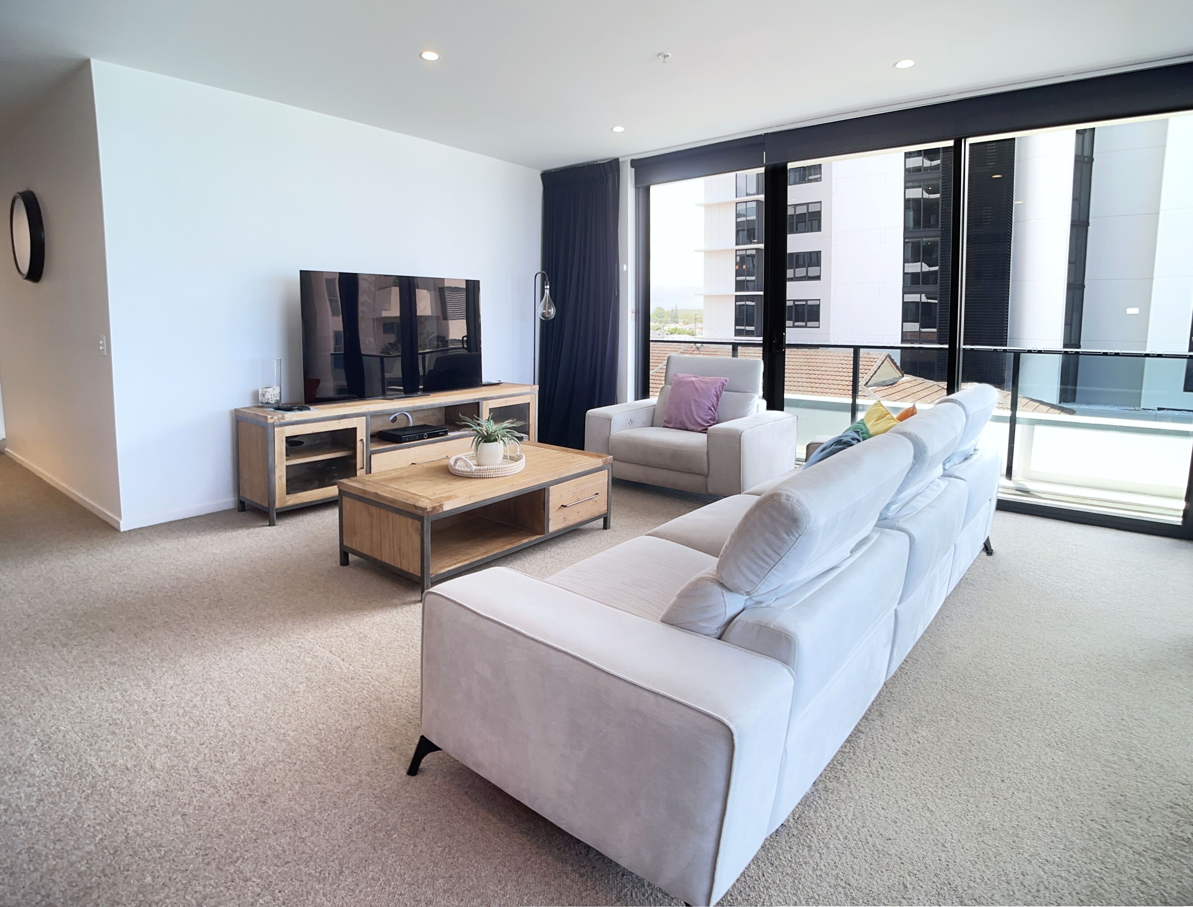 A modern living room with a white sofa, a wooden coffee table, a TV, and a balcony with a view of buildings.