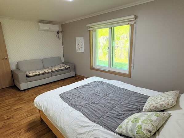 A bedroom with a bed, a grey sofa, a window with a view, and an air conditioning unit.