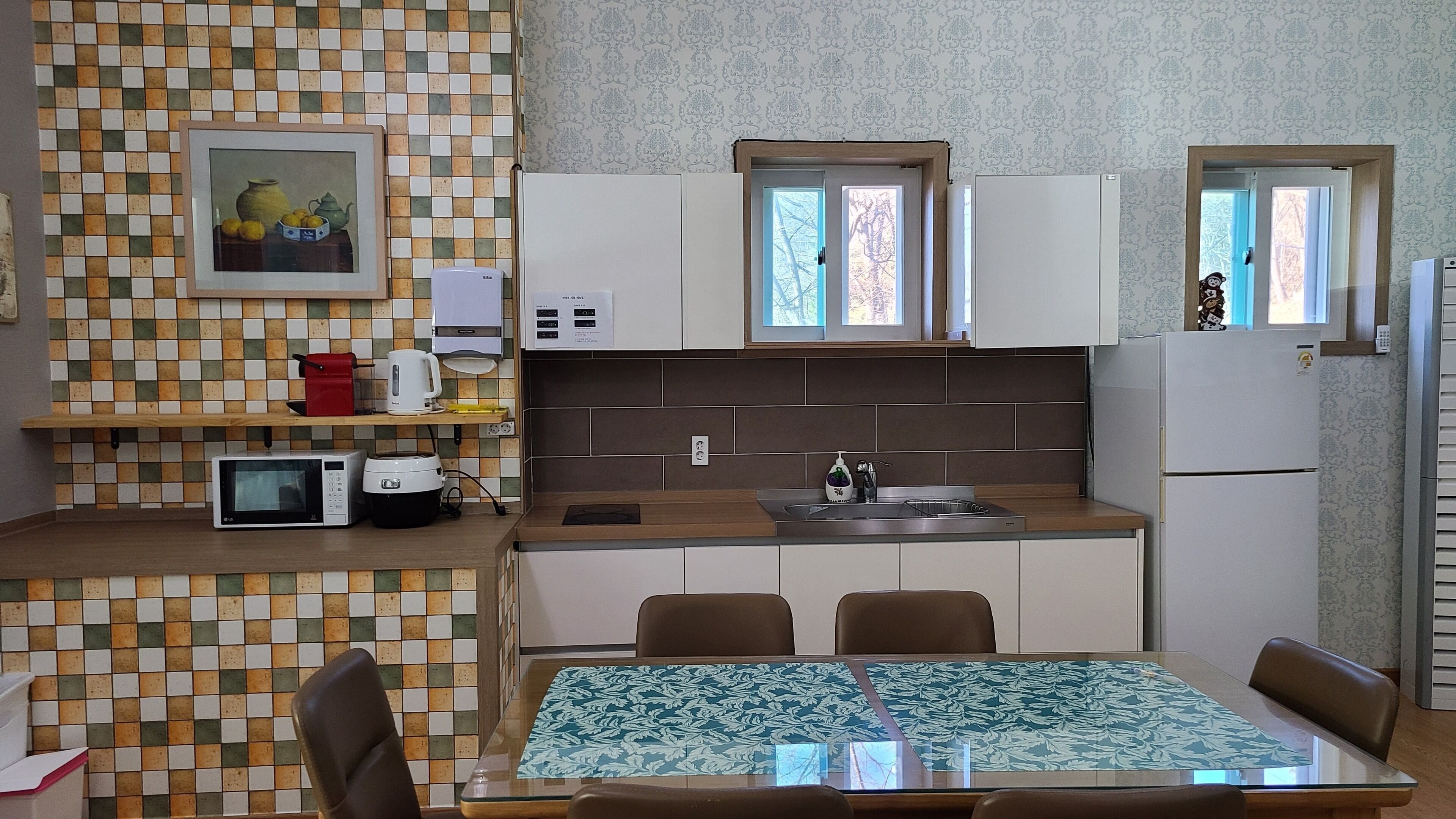 A compact kitchen with a microwave, kettle, and a sink. There is a dining table with chairs and a patterned tablecloth.