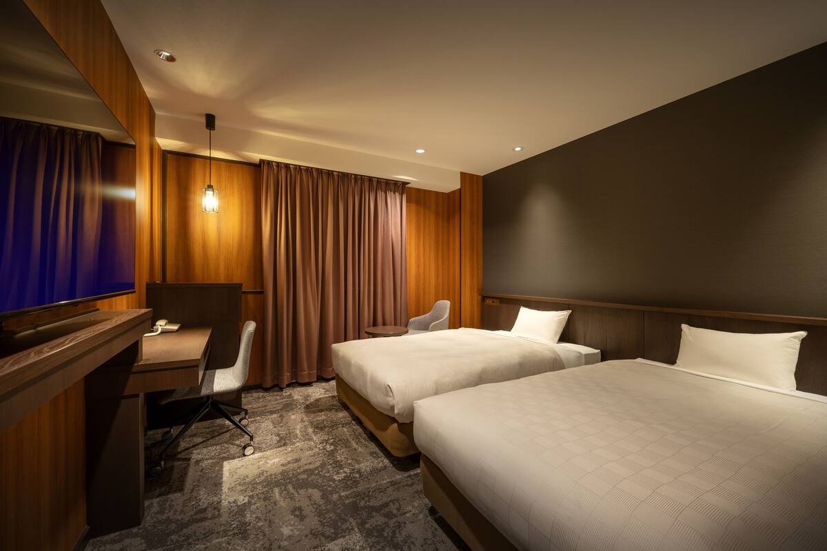A hotel room with two beds, a desk with a chair, a television, and a large window with curtains.