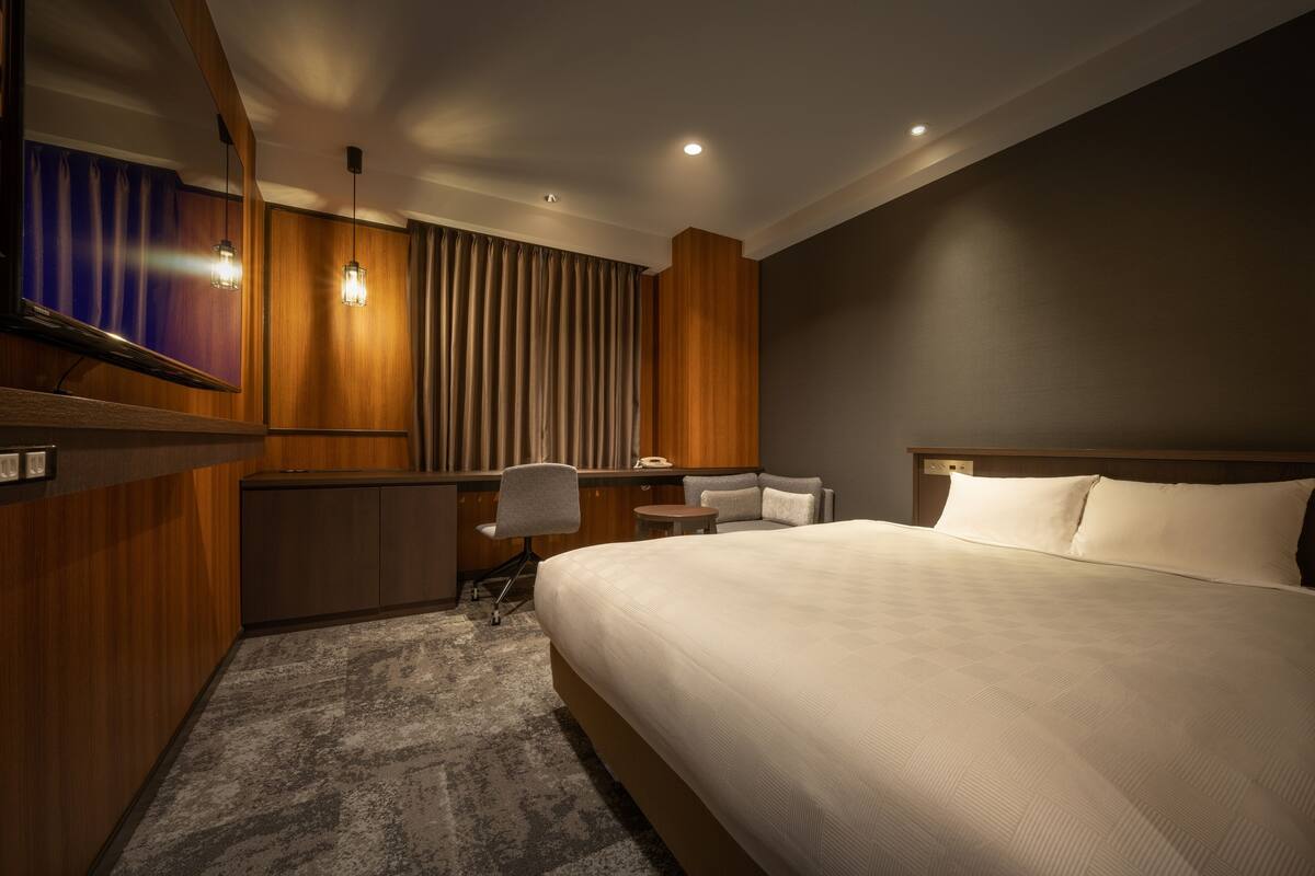 A modern hotel room with a large bed, a desk with a chair, a small table, and a television.