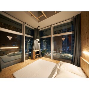 Ein modernes Schlafzimmer mit einem großen Bett, einem Nachttisch und Blick auf eine Stadtlandschaft durch raumhohe Fenster.
