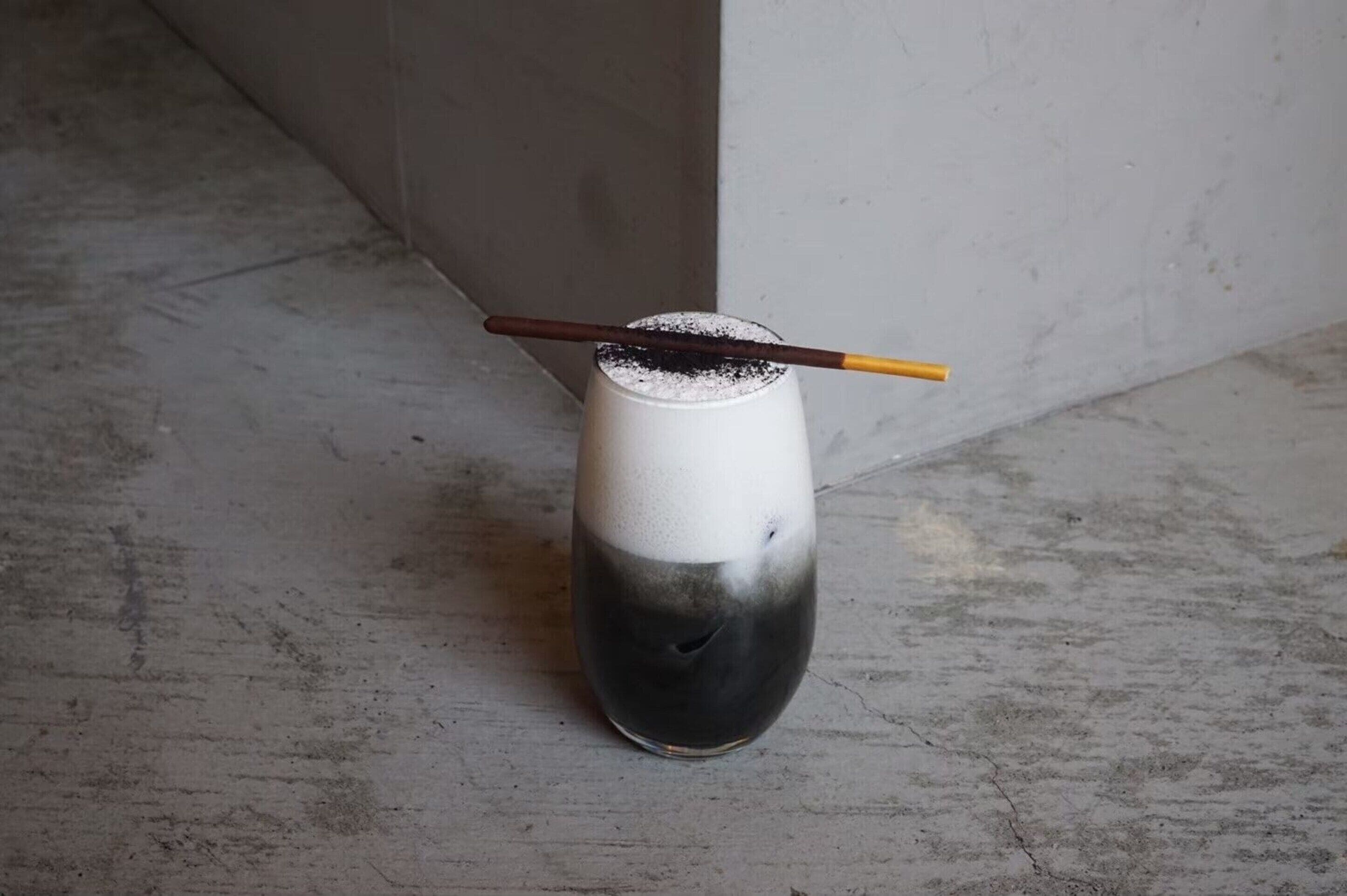 A glass with a layered drink and a straw, placed on a concrete floor.