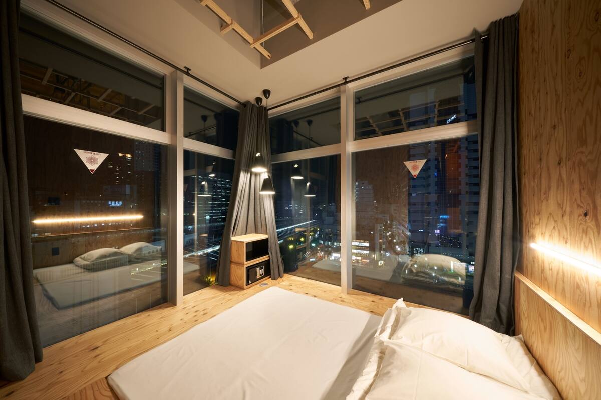 A modern bedroom with a large bed, wooden headboard, and a city view through floor-to-ceiling windows.