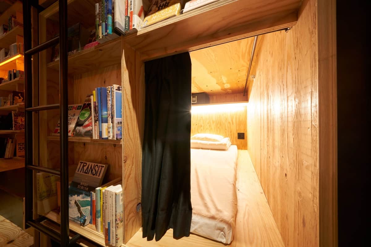 A small, cozy room with a wooden interior, a bookshelf filled with books, a single bed with white bedding, and a black curtain partially drawn.