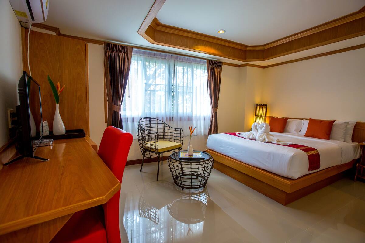 A hotel room with a wooden desk, a red chair, a bed with white bedding and orange pillows, a small round table with a vase, and a window with curtains.